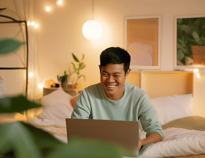 An Indonesian man, seated in a warmly lit bedroom, smiles at his laptop screen, captivated by the content displayed before him.